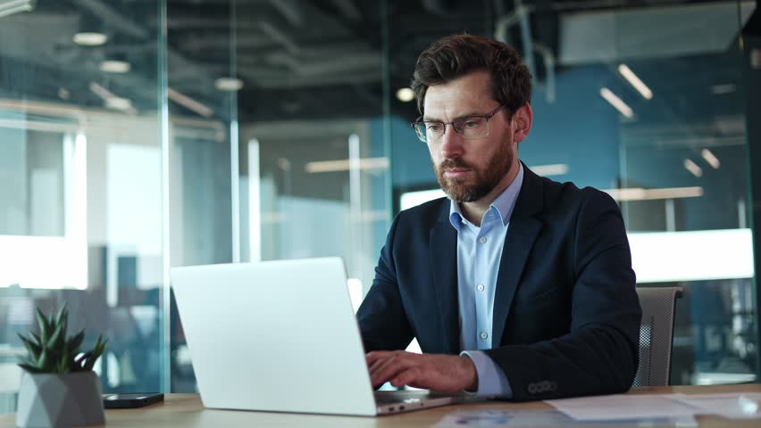 Portrait of handsome male wearing protective glasses for working at personal laptop in cabinet interior. Successful man sitting in front of modern gadget and actively engaging in work process indoors.