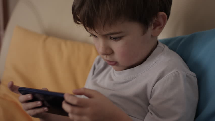 Little Boy Gamer Playing Video Game on Mobile Phone. Child Happy Playing Games With Phone. Preschooler Kid Plays Video Game On Smartphone. Harm for Mental Kid Health Eyesight Gambling Addiction.