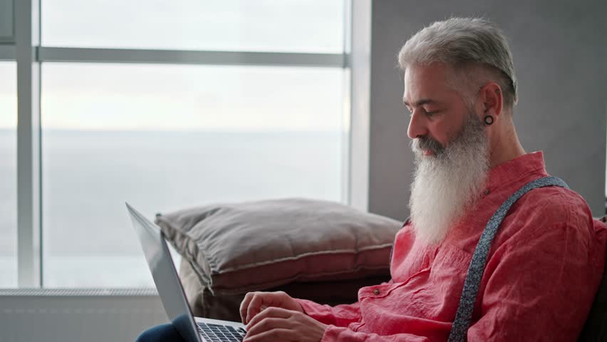 Side view of an elderly man with gray hair and a full beard with a ring in his ear and a pink shirt sitting on a modern sofa and typing on his gray laptop in a modern apartment overlooking the sea