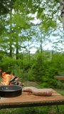 Vertical video of female hands cutting raw sausage rings or freshly stuffed sausage on wooden board near bonfire and spring forest. Preparing breakfast on camping. Food, travel and wanderlust concept