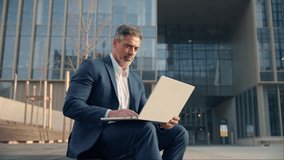 Focused mature successful businessman working, scrolling, typing on laptop sitting outdoors at office building. Handsome older man entrepreneur in suit using wireless pc computer for online business - Powered by Shutterstock - Get 15% off with code: PIKWIZARD15