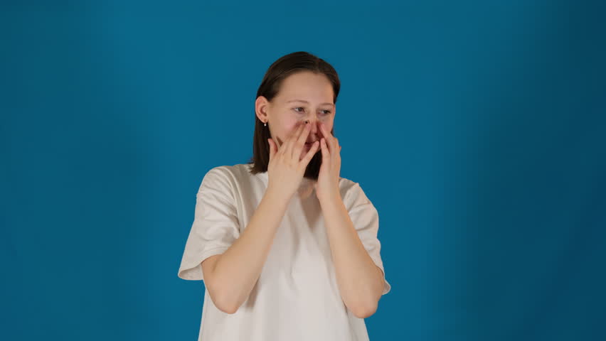 Sad woman wipes tears from face on blue background. Young lady stands showing facial expression with mixture of sorrow and resilience Royalty-Free Stock Footage #3482571735