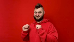 Horizontal 4K video with copyspace. Bearded adult joyful man with mohawk in scarlet hoodie looks at camera, smiles. Young man with beard dances on red background. Concept of monochrome, joy, holiday