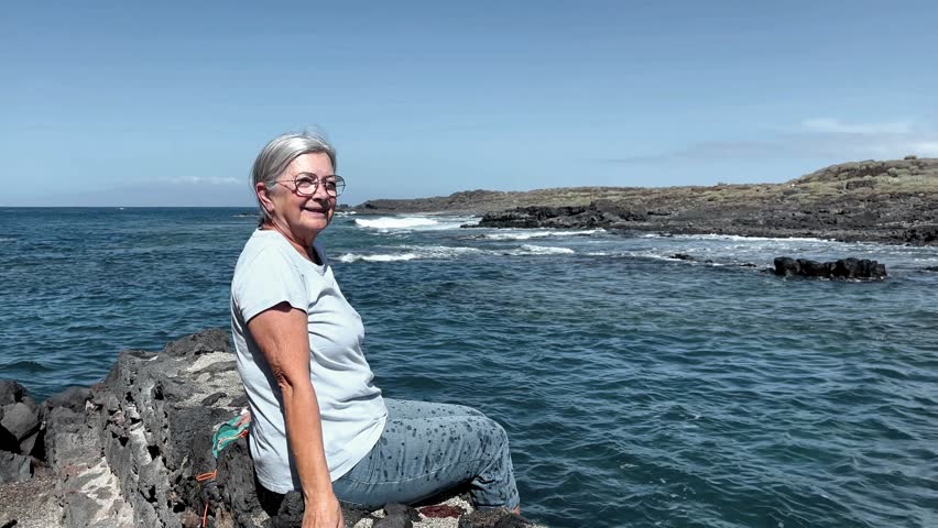 Smiling senior white-haired woman sitting on a rocky beach looking the waves crashing on the rocks. Elderly female in retirement or vacation enjoying a sunny day and freedom outdoors