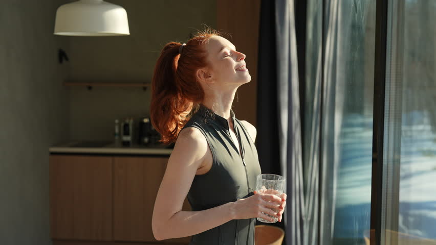 Young athletic woman in sport outfit standing by window and drinks fresh water in morning after fitness training workout exercise at home, enjoying sunny weather and lifestyle, taking care of health.