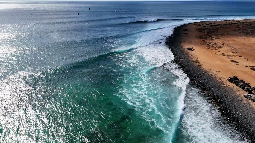 Flying over the coastline of Ponta Preta, Sal. A big wave breaks. Kiters and surfers can be seen
