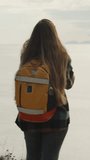 Vertical video. View from Behind of a Young Solo Female Traveler with a Backpack on the Edge of a Cliff by the Sea. She has Folded her Hands.