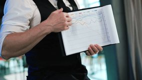 Unrecognized man in white shirt and black vest holding a piece of paper with graphs. Man shows thumb up displaying charts. Close up. - Powered by Shutterstock - Get 15% off with code: PIKWIZARD15