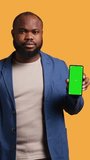 Vertical video Smiling african american man presenting cellphone with green screen display, isolated over studio background. Cheerful BIPOC person creating promotion with blank copy space mockup phone