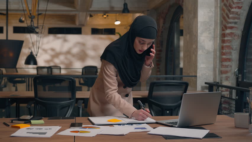 Muslim islamic Arabian woman girl in hijab working laptop project analysis planning female smile businesswoman talking mobile phone stand at table write notes speak smartphone business call in office - Powered by Shutterstock - Get 15% off with code: PIKWIZARD15