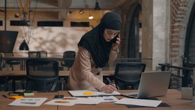 Muslim islamic Arabian woman girl in hijab working laptop project analysis planning female smile businesswoman talking mobile phone stand at table write notes speak smartphone business call in office - Powered by Shutterstock - Get 15% off with code: PIKWIZARD15