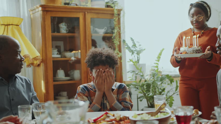 Little African American boy covering his eyes, then blowing candles on cake and smiling while family clapping hands and greeting him on Birthday dinner party at home