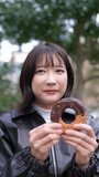 Vertical video of a young Japanese woman in her 20s sitting and eating donuts in a park around Gotanda, Shinagawa-ku, Tokyo in winter