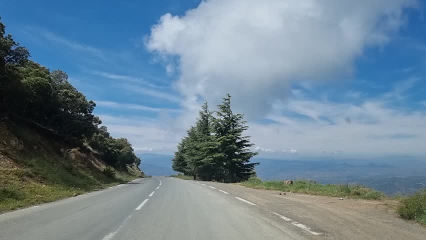 As the winding road gently weaves through the majestic Algerian mountains, the dramatic clouds in the sky create a breathtaking backdrop for a scenic journey