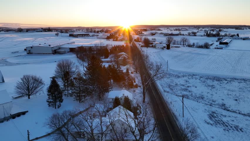 Golden hour sunrise over snow covered landscape in rural USA. Aerial truck shot over road with houses and fields.