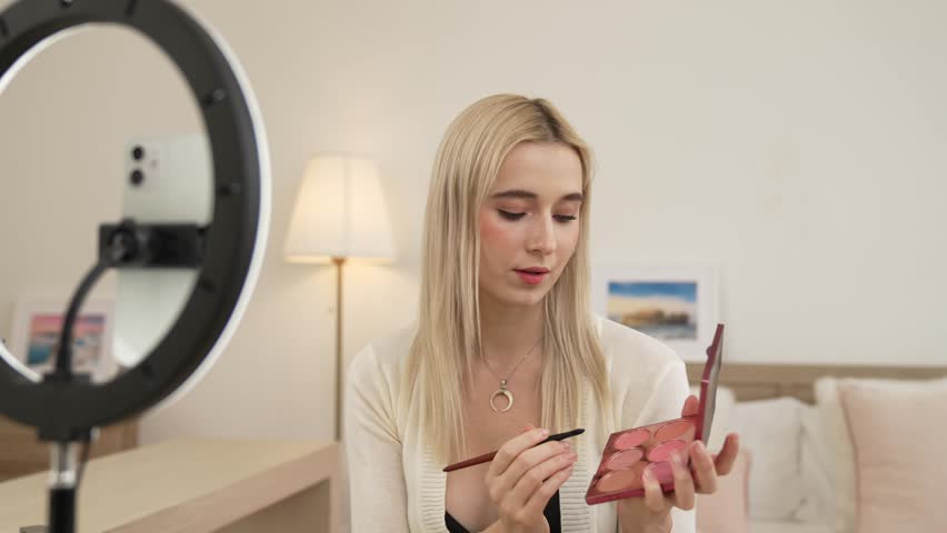 Young woman making beauty and cosmetic tutorial video content for social media. Beauty blogger using camera and light ring while showing how to apply eyeshadow to audience or follower. Blithe Royalty-Free Stock Footage #3483113751
