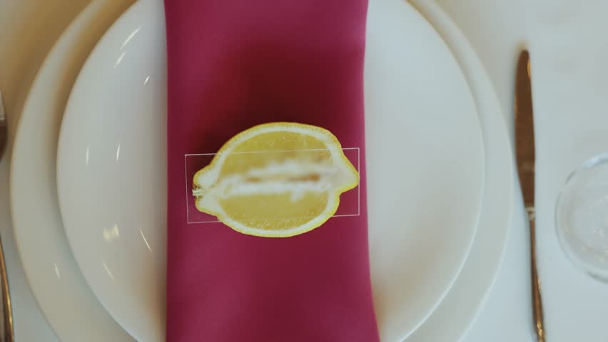 A magenta color napkin with a lemon slice sits on a plate on the table, as a wedding serve, white classic plate and golden color fork and knife laying near, slow motion.