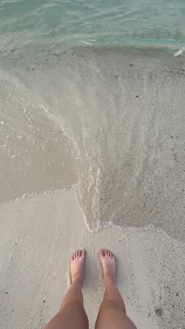 Closeup top view video of legs of woman enjoying summer vacations standing on sandy sea beach. Sea waves wash the woman