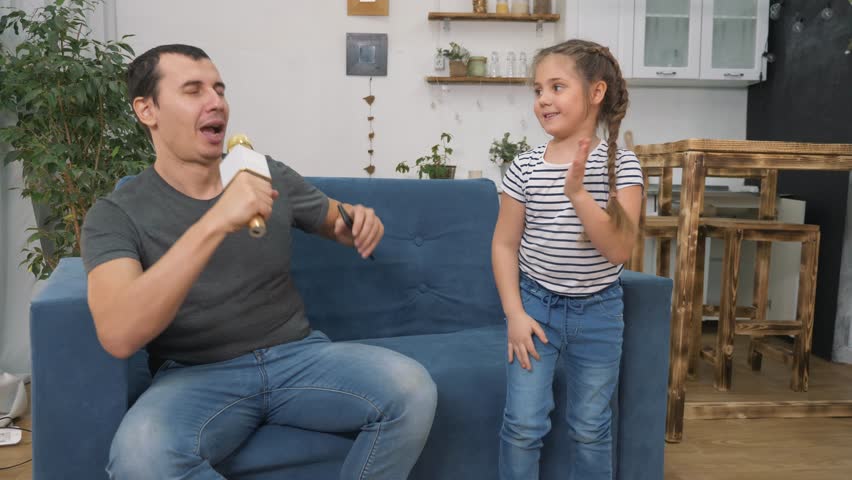 Happy family concept.dad and child spend time together at home on the sofa. father's day happy father sings songs loudly into microphone.little girl covers her ears first from loud music after dancing Royalty-Free Stock Footage #3483258143