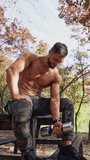 Shirtless sportsman doing workout in autumn park. Active man with muscular body sitting on a bench and lifting dumbbell on nature background. Vertical video
