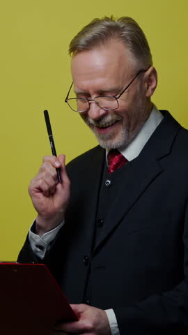 Happy elderly businessman with folder. Grey-bearded senior man is writing on paper on yellow background. Positive emotions. Vertical video