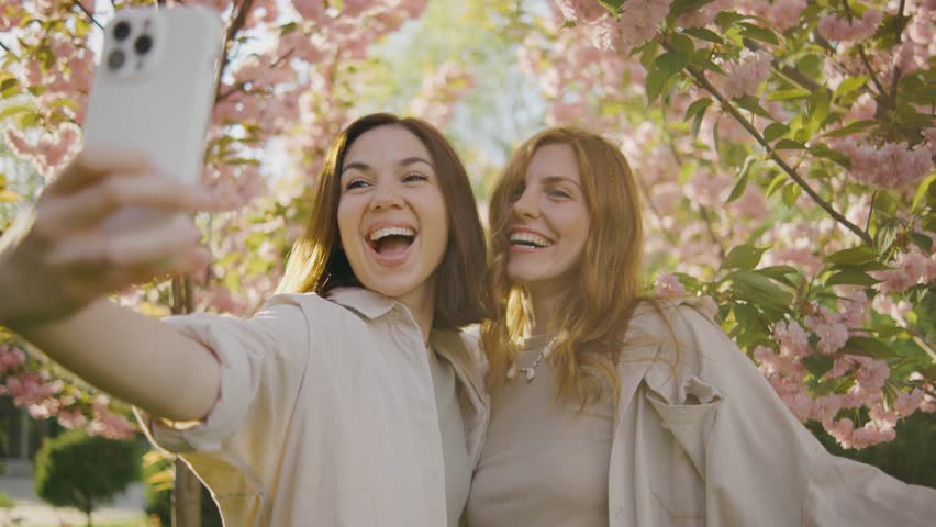 two incredibly beautiful young women take a selfie on a smartphone in a park against the background of pink blooming spring trees. Girls laugh, rejoice and make faces at the camera