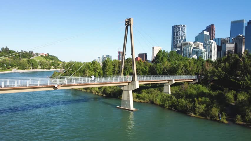 Downtown Calgary, Alberta, Canada skyline. Wide aerial drone view. Panorama from the walking bridge over the Bow River on a sunny blue skies day. Prince Island Park. Royalty-Free Stock Footage #3483490359