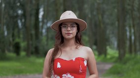 Ecuadorian woman in a vibrant red outfit, adorned with glasses and a cowboy hat, stands amidst the forest backdrop, forming a heart shape with her hands and sharing a radiant smile. - Powered by Shutterstock - Get 15% off with code: PIKWIZARD15