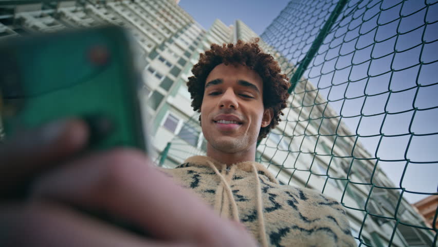 Happy zoomer chatting phone surrounded by cityscape closeup bottom view. Carefree young man smiling reading message on smartphone at street. Curly hispanic guy browsing social media at city district.