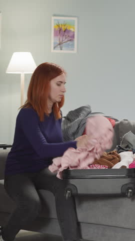 Angry redhead woman collects things after conflict with husband. Disappointed wife puts clothes into suitcase sitting on sofa in living room slow motion Royalty-Free Stock Footage #3483669511