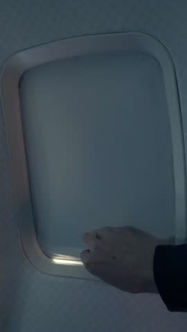 A vertical dolly view of an airplane passenger raising a window shade to reveal a white bloom background. Great opening shot to reveal a travel montage.  	