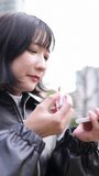 Vertical video of a young Japanese woman in her 20s sitting and putting on makeup in a park around Gotanda Station, Shinagawa-ku, Tokyo in winter