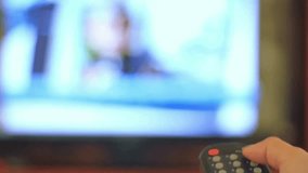 Male hand holding the TV remote control. Channel surfing, focused on the hand and remote control. Close up view. Blurry tv at the background. Person controls TV using remote control.