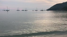 video clip of man wears orange short pants sailing on white surfboard in scene of beautiful beach when sunset with clear water, many yachts in the sea and amazing wave, taken by smart phone