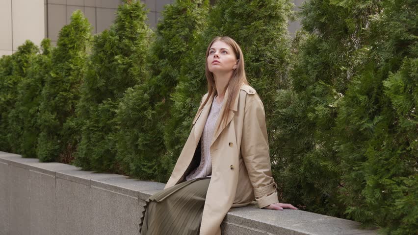 Three quarters shot of a young woman in a light coat sitting on a parapet by green thujas and then jumping off.