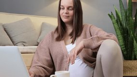 Happy pregnant woman making video call on laptop while relaxing on floor near sofa having pleasant conversation waving hand greeting gesture smiling to laptop screen - Powered by Shutterstock - Get 15% off with code: PIKWIZARD15