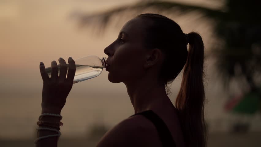 Woman drinking water after training. Fitness thirsty girl drinks water during workout pause, after a grueling run of a long sea ocean beach at sunset. Endurance sport, fitness, morning drink concept.