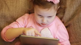 A little cute girl is sitting in sofa in a living room, looking cartoon and playing the game on a tablet.. Funny child looks on the computer screen and plays downloaded application. Close-up portrait