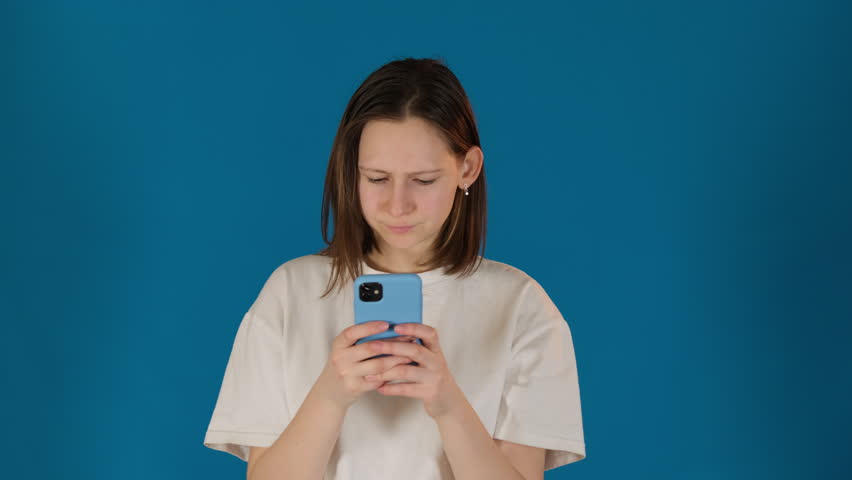Concentrated young woman types message on smartphone. Lady in t-shirt intensely responds in online chat on blue background