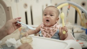 A baby is eating from a high chair with a spoon in his mouth. The baby is wearing a pink bib and a pink bandana. The baby is smiling and seems to be enjoying his meal - Powered by Shutterstock - Get 15% off with code: PIKWIZARD15
