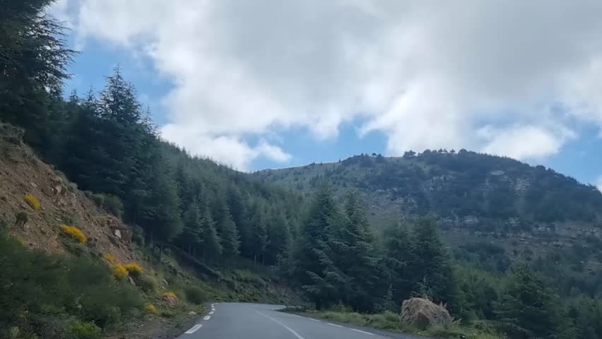 As the winding road gently weaves through the majestic Algerian mountains, the dramatic clouds in the sky create a breathtaking backdrop for a scenic journey