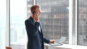 Busy professional business man manager, young male executive, businessman entrepreneur using laptop computer talking on phone making call working at office standing near big skyscraper window. - Powered by Shutterstock - Get 15% off with code: PIKWIZARD15