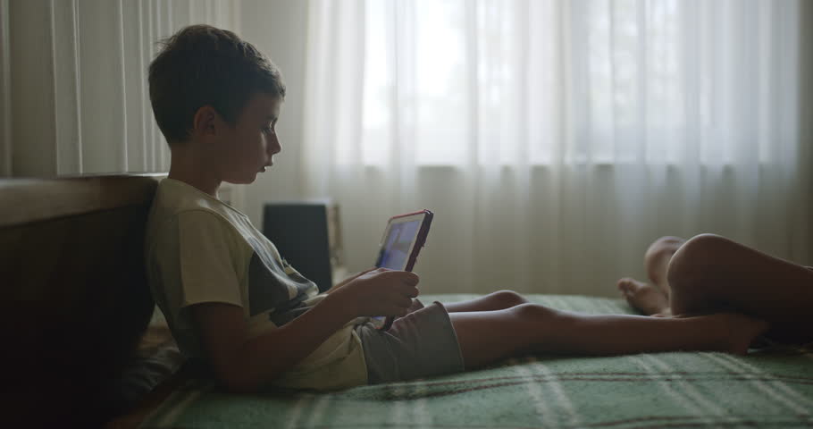 A boy rests on a bed engrossed in a digital tablet, with a younger child lying next to him in a serene, sunlit bedroom.
