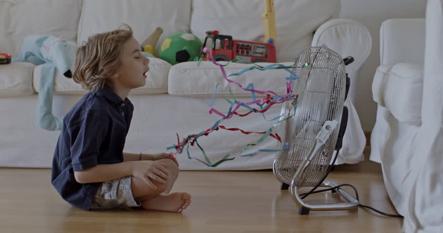 A happy, young boy with blond hair is seated indoors, extending his hand to feel the breeze from a standing electric fan, with toys scattered in the background.
