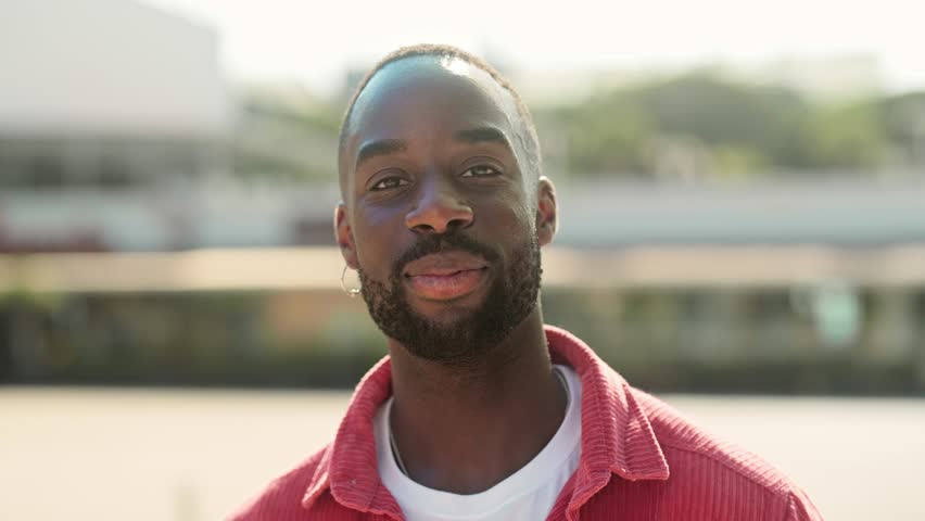 Smiling cool generation z Black hipster guy wearing red shirt posing outdoors. Close up portrait of happy young adult African man hipster standing on city street looking at camera.
