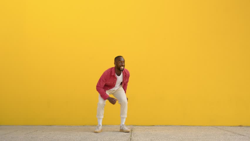 Happy active energetic crazy gen z young Black man jumping at yellow urban wall. Excited cool funky African guy having fun dancing moving on background celebrating victory. Full body video.