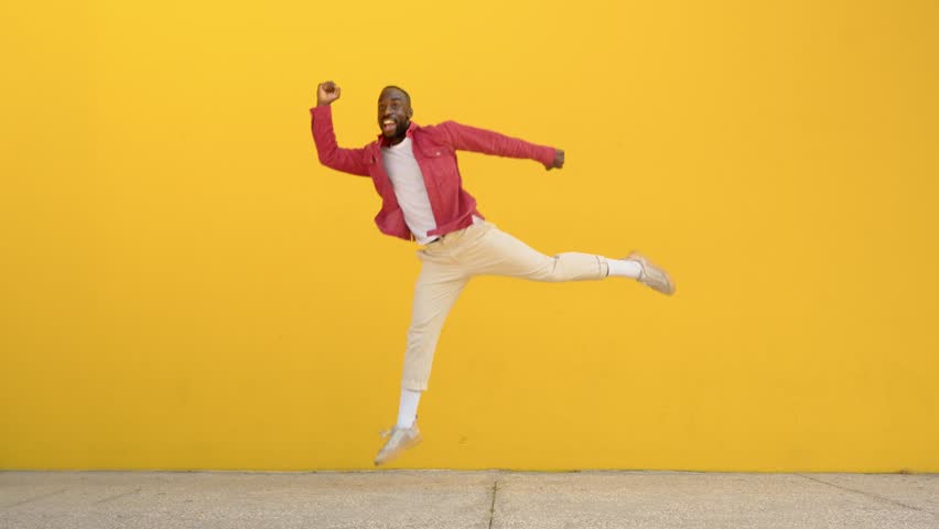 Happy active energetic crazy gen z young Black man jumping at yellow urban wall. Excited cool funky African guy having fun dancing moving on background celebrating victory. Full body video.