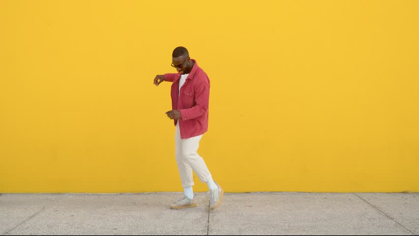 Happy excited cool stylish funky young African Black man dancer wearing sunglasses feeling joy having fun dancing, jumping standing at yellow color urban wall background. Full body.