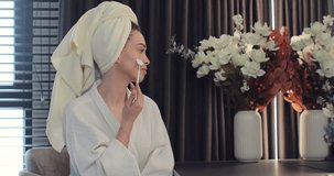 Young woman wearing bathrobe and towel on head doing makeup using brush in front of mirror in bedroom. Enjoying natural beauty in morning routine.