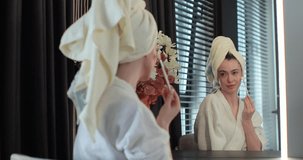 Young woman wearing bathrobe and towel on head doing makeup using brush in front of mirror in bedroom. Enjoying natural beauty in morning routine.